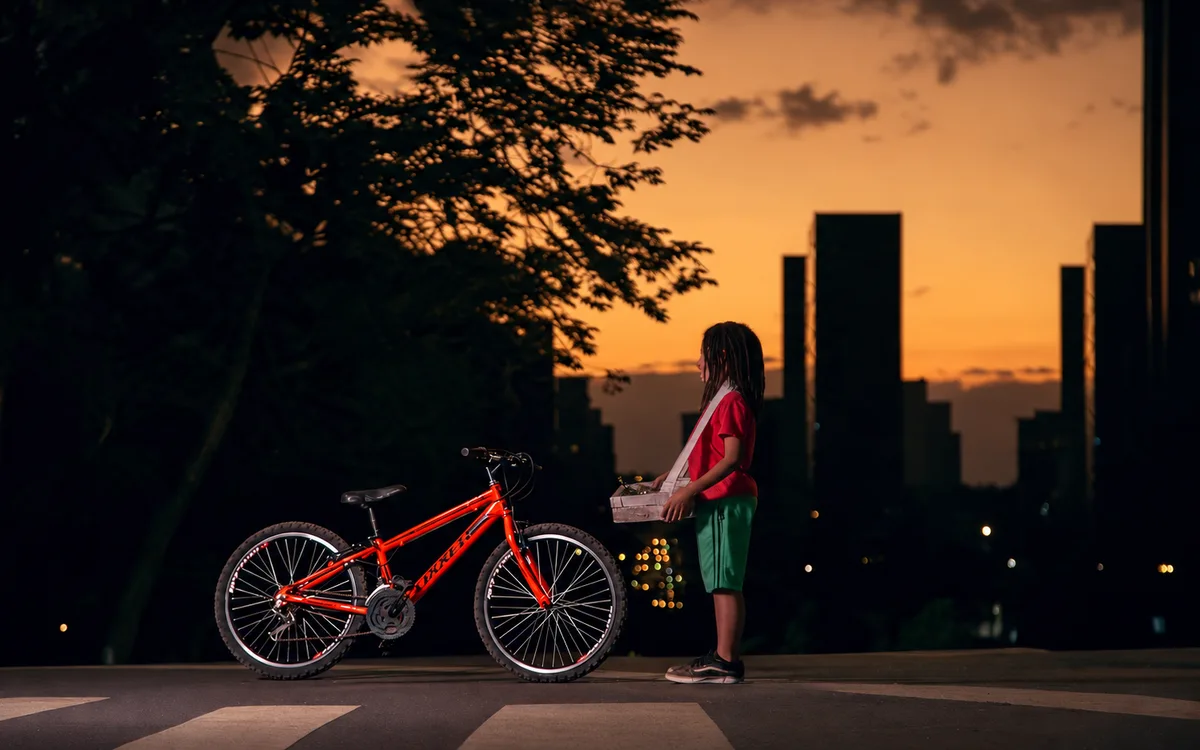 Pedro, de camiseta vermelha, com sua bicicleta vermelha diante da cidade ao pôr do sol.