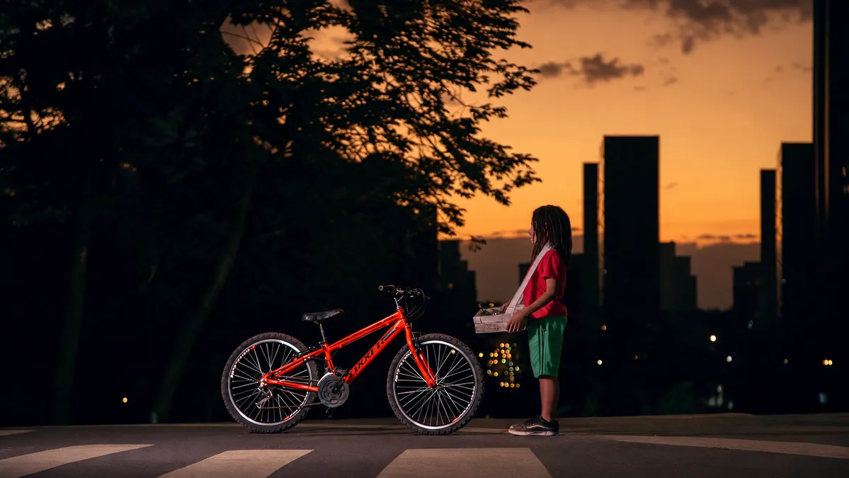 Pedro, de camiseta vermelha, com sua bicicleta vermelha diante da cidade ao pôr do sol.